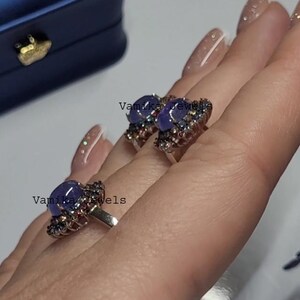 May include: Two silver rings with a large, oval, blue-purple gemstone at the center. The gemstones are surrounded by smaller, colorful stones. The rings are displayed on a hand. The text "Vamika Jewels" is visible.