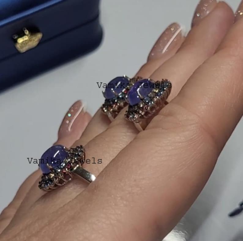 May include: Two silver rings with a large, oval, blue-purple gemstone at the center. The gemstones are surrounded by smaller, colorful stones. The rings are displayed on a hand. The text "Vamika Jewels" is visible.