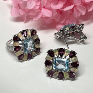 May include: A silver ring and matching earrings set. Each piece features a rectangular, light blue gemstone surrounded by pear-shaped, dark red and light yellow gemstones. The earrings have a post back.