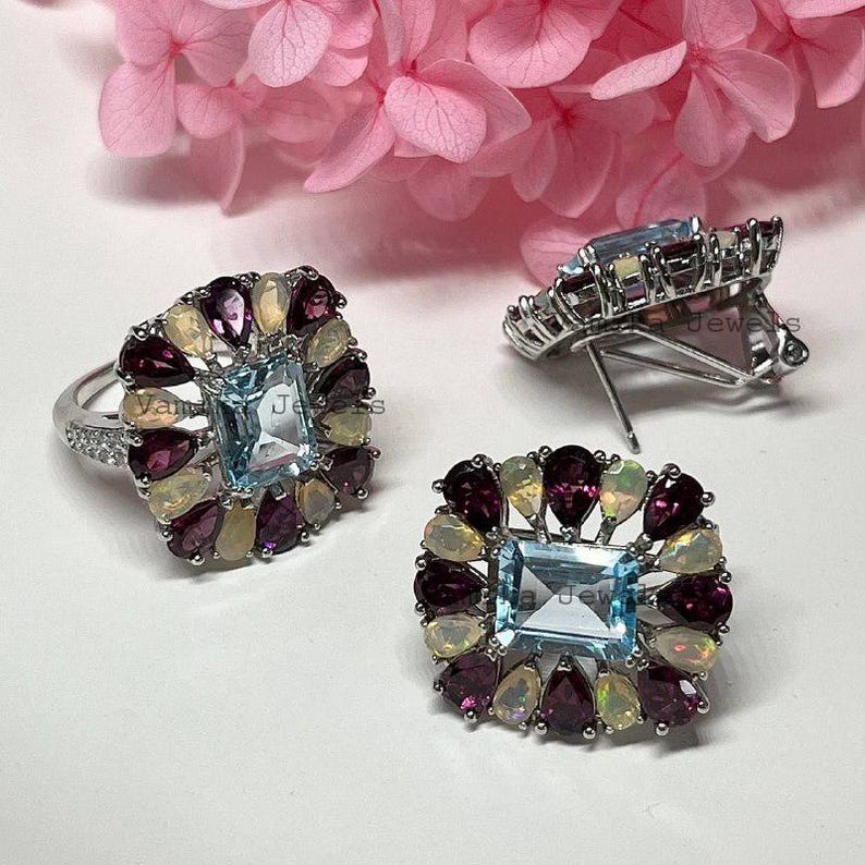 May include: A silver ring and matching earrings set. Each piece features a rectangular, light blue gemstone surrounded by pear-shaped, dark red and light yellow gemstones. The earrings have a post back.