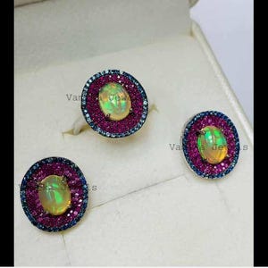 May include: A jewelry set featuring an oval opal gemstone surrounded by a ring of small, pink gemstones and a border of blue gemstones. The set includes a ring and two matching earrings. The text "Vara Jewels" is visible.