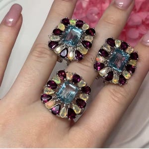 May include: Three silver rings with a square, light blue gemstone center, surrounded by a halo of smaller, teardrop-shaped gemstones in shades of purple, yellow, and white. The rings are displayed on a person's hand.