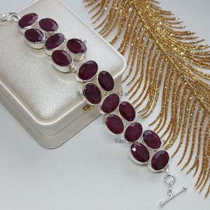 Puede incluir: Una pulsera de plata con piedras preciosas ovaladas de color rojo intenso. Las piedras están engastadas en biseles individuales de plata y unidas entre sí. La pulsera se exhibe sobre una superficie blanca con un elemento decorativo dorado en el fondo.