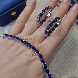 May include: A jewelry set featuring a bracelet and two hoop earrings. The bracelet and earrings are adorned with oval-cut, deep blue gemstones set in a silver-toned metal. The jewelry is displayed on a hand, with a blue jewelry box in the background.
