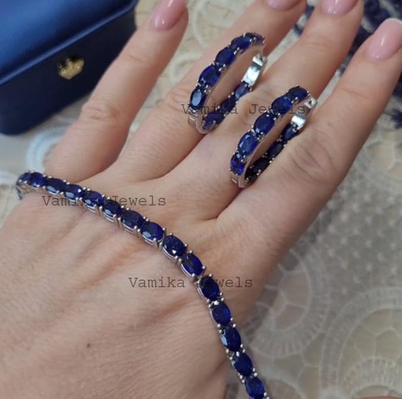 May include: A jewelry set featuring a bracelet and two hoop earrings. The bracelet and earrings are adorned with oval-cut, deep blue gemstones set in a silver-toned metal. The jewelry is displayed on a hand, with a blue jewelry box in the background.