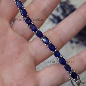 May include: A silver bracelet with oval, deep blue gemstones. The bracelet is clasped and features the text "Vamika Jewels". The gemstones are set closely together, creating a continuous line of color. The bracelet is shown against a blurred background.