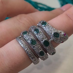 May include: Four silver snake rings with emerald green stones and small diamonds. The rings are stacked on a finger.