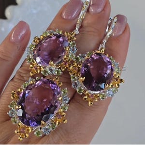 May include: A jewelry set featuring a large oval amethyst gemstone pendant and matching earrings. The amethyst is surrounded by smaller gemstones in shades of green, pink, and blue, set in a gold and silver setting.
