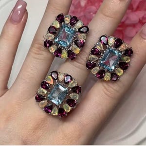May include: Three square-shaped rings with a central light blue rectangular gemstone, surrounded by smaller gemstones in shades of purple, yellow, and white. The rings are set on a silver band.