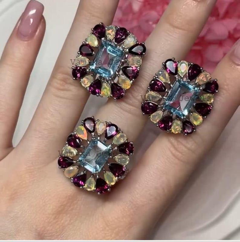May include: Three square-shaped rings with a central light blue rectangular gemstone, surrounded by smaller gemstones in shades of purple, yellow, and white. The rings are set on a silver band.