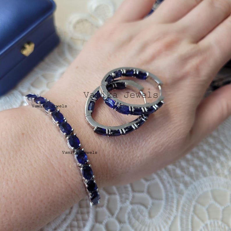 May include: A silver bracelet and hoop earrings set with oval, dark blue gemstones. The jewelry is displayed on a person's hand, with a blue jewelry box in the background. The earrings are hoop style.