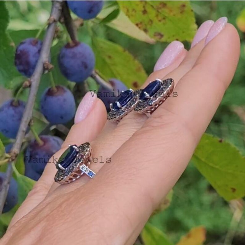 May include: A silver ring and earrings set featuring a large blue gemstone surrounded by smaller black gemstones. The jewellery is displayed on a hand against a background of green leaves and dark blue berries.