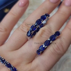 May include: Close-up of two rings and a bracelet featuring oval-cut, deep blue gemstones. The jewelry is set in a silver or white metal. The rings are on a person's finger. The brand name "Vamika Jewels" is visible.