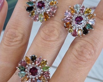 May include: A jewelry set consisting of two stud earrings and one ring, each featuring a central round dark pink gemstone surrounded by a halo of multi-colored pear-shaped stones in shades of yellow, green, pink, and clear set in silver.