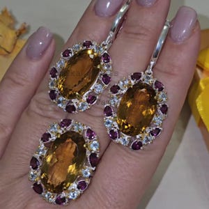 May include: A jewelry set featuring a ring and a pair of earrings. Each piece has a large, oval, amber-colored stone surrounded by smaller, round, purple and light blue stones, set in silver metal.