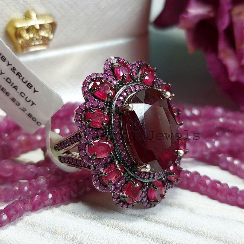 May include: An elaborate ring with a central oval ruby, encircled by smaller rubies in a floral pattern. The ring has a silver band. The image shows pink beads and a rose in the background.