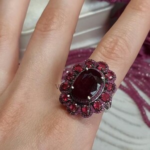 May include: A striking ruby ring. The ring features a large, oval-cut, dark red ruby at its centre, encircled by a halo of smaller, lighter red rubies. The setting is a light-colored metal, possibly silver.