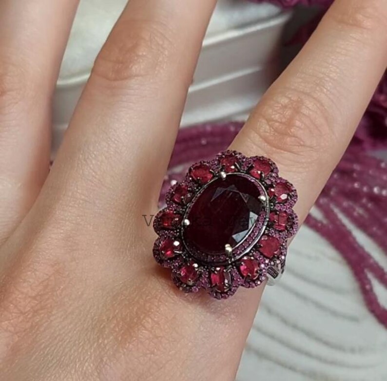 May include: A striking ruby ring. The ring features a large, oval-cut, dark red ruby at its centre, encircled by a halo of smaller, lighter red rubies. The setting is a light-colored metal, possibly silver.