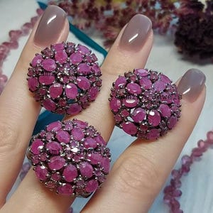 Puede incluir: Tres anillos con un grupo de piedras preciosas rosas oscuras de corte ovalado. Los anillos están colocados en una mano con uñas pulidas. Los anillos son de Yamika Jewels.