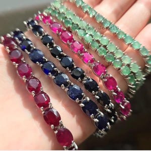 Könnte beinhalten: Eine Auswahl an Armbändern mit Edelsteinen in Rubinrot, Saphirblau, Smaragdgrün und Pink. Jedes Armband hat ovale Steine, die in einem silberfarbenen Metall gefasst sind.
