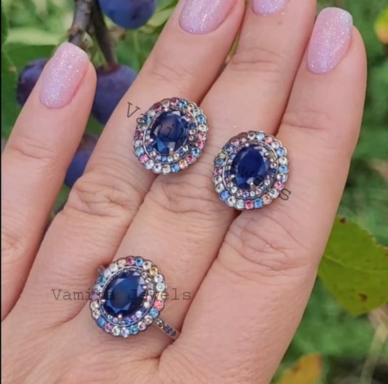 May include: A set of three silver rings featuring blue oval gemstones encircled by a halo of colourful gemstones. The rings are displayed on a hand with pink nail polish.
