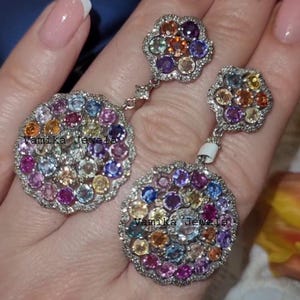 May include: A pair of silver earrings featuring a floral design with a cluster of colorful gemstones. The earrings have a round, dangling pendant with a variety of colored stones, including pink, blue, and orange. The text "Vamika Jewels" is visible.
