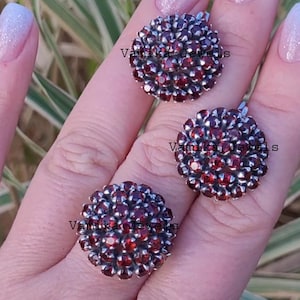 Può includere: Sono esposti tre gioielli, tra cui due orecchini e un anello. Ogni pezzo presenta un gruppo di piccole pietre rotonde, rosso scuro, incastonate in metallo argentato. Il testo "Vanika Jewels" è visibile su ogni pezzo.