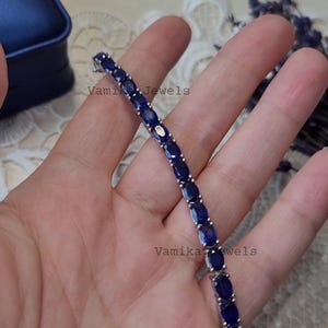 May include: A silver bracelet featuring oval-cut, deep blue gemstones. The gemstones are set closely together, creating a continuous line of color. The bracelet is displayed on a light background. The text "Vamika Jewels" is visible.