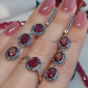 Op de afbeelding: Een sieradenset met een ring en oorbellen. De ring en oorbellen hebben ovale, dieprode edelstenen omringd door kleine blauwe stenen. De oorbellen zijn lang en bungelend. De sieraden zijn zilverkleurig.