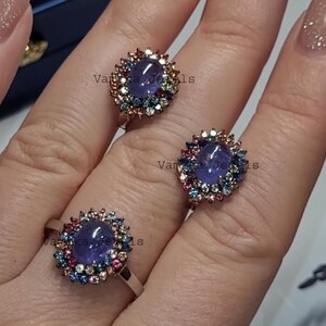 May include: Three silver rings with a central oval, purplish-blue gemstone, surrounded by a halo of smaller, colorful gemstones. The rings are displayed on a light background.