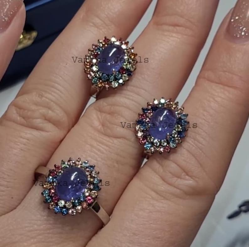 May include: Three silver rings with a central oval, purplish-blue gemstone, surrounded by a halo of smaller, colorful gemstones. The rings are displayed on a light background.