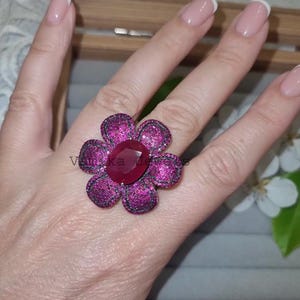 Pu&ograve; includere: Un anello vistoso a forma di fiore, con un rubino ovale centrale e petali con piccole pietre rosa e nere scintillanti. L'anello &egrave; indossato su una mano con unghie curate. Il testo "Vantika Jewels" &egrave; visibile.