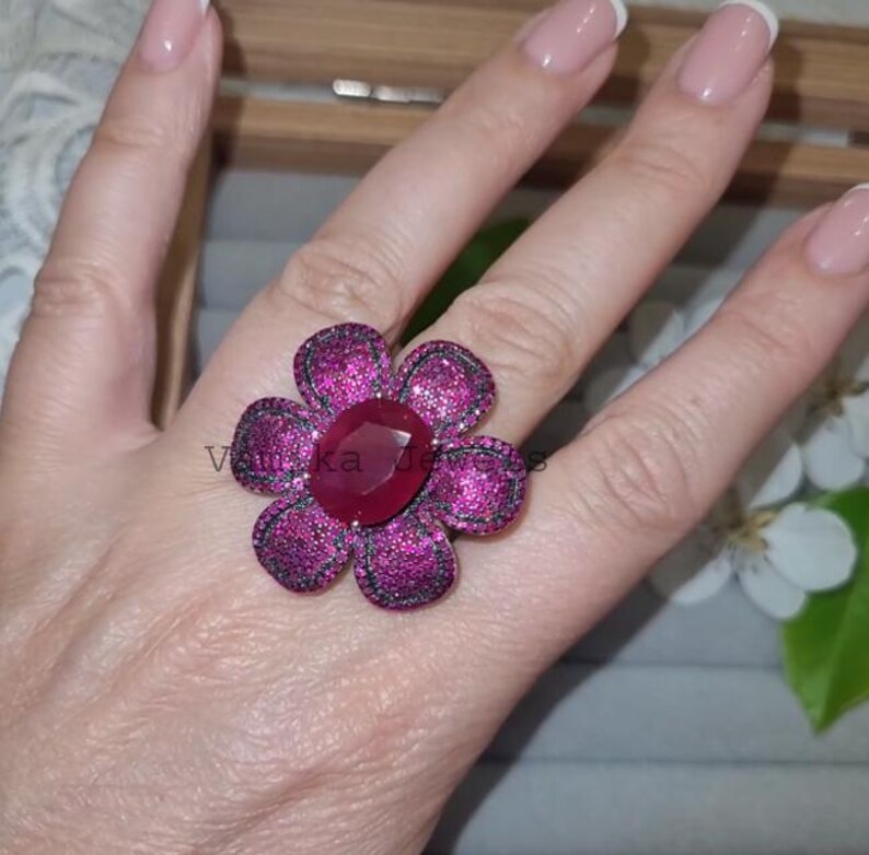 Pu&ograve; includere: Un anello vistoso a forma di fiore, con un rubino ovale centrale e petali con piccole pietre rosa e nere scintillanti. L'anello &egrave; indossato su una mano con unghie curate. Il testo "Vantika Jewels" &egrave; visibile.