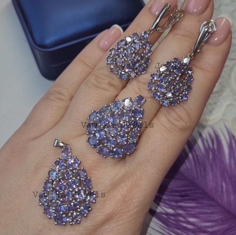 May include: A jewellery set comprising a pendant, ring, and earrings. Each item is embellished with clusters of small, oval, light purple gemstones. The metal setting is silver-toned, and the gemstones are arranged in a teardrop shape.