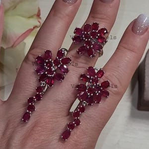 Op de afbeelding: Een sieradenset met een ring en oorbellen. De ring en oorbellen zijn zilverkleurig en versierd met clusters van ovale, dieprode edelstenen in een bloemmotief. De oorbellen hebben een trapsgewijs ontwerp.