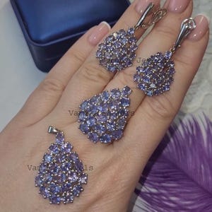 May include: A jewellery set comprising a ring, pendant, and earrings, each piece set with clusters of small, light purple gemstones. The earrings are drop-style, and the ring has a large, oval cluster. The pendant is teardrop-shaped.