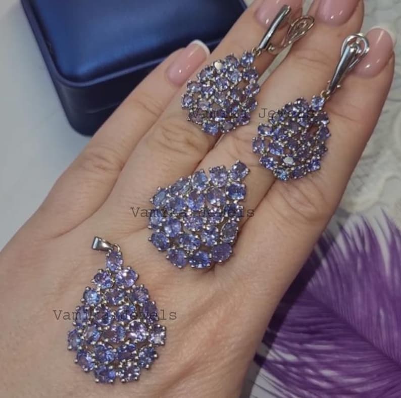 May include: A jewellery set comprising a ring, pendant, and earrings, each piece set with clusters of small, light purple gemstones. The earrings are drop-style, and the ring has a large, oval cluster. The pendant is teardrop-shaped.