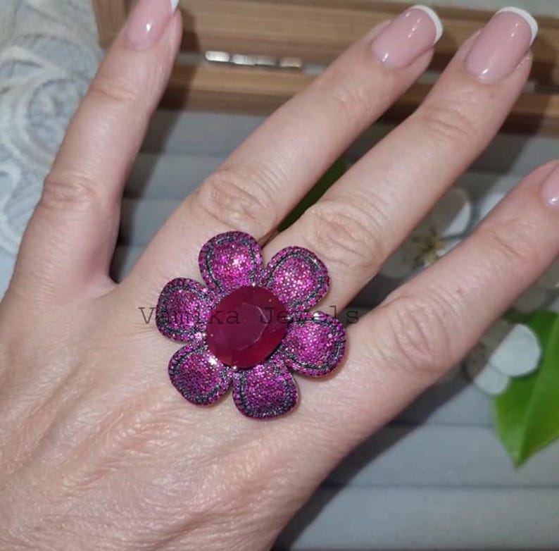 Pu&ograve; includere: Un anello a forma di fiore di grandi dimensioni con una pietra di rubino ovale al centro. I petali sono incastonati con pietre rosa pi&ugrave; piccole e scintillanti e la fascia dell'anello &egrave; argentata. L'anello &egrave; su una mano con unghie curate.