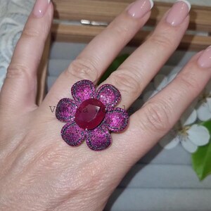 Pu&ograve; includere: Un anello a forma di fiore magenta con una grande pietra centrale ovale rosso rubino. I petali sono magenta con dettagli neri. L'anello &egrave; su una mano. Il testo "Vinka Jewels" &egrave; visibile sull'anello.