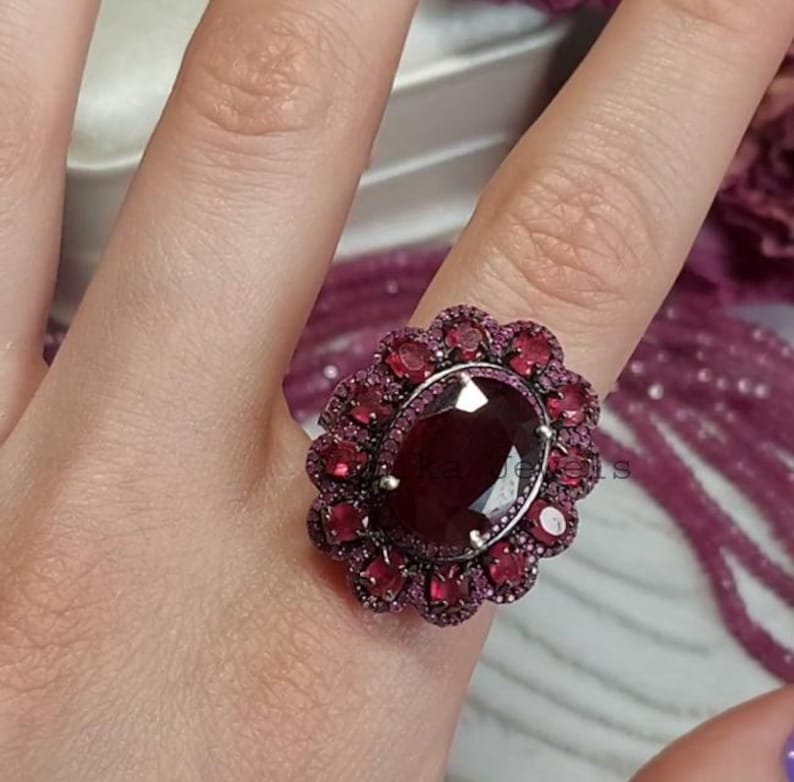 May include: An elaborate ring with a large, oval-cut, deep red gemstone at its centre, encircled by smaller red stones. The ring has a floral design and a silver-toned band. The gemstones are cut to reflect light.