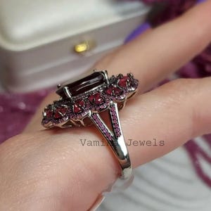 May include: A silver ring with a large, rectangular, dark red gemstone at the centre, encircled by smaller, round, red gemstones. The ring's band has a split design with more small red gemstones. The text "Vami Jewels" is visible.