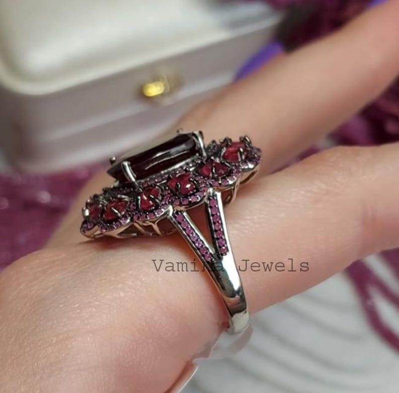 May include: A silver ring with a large, rectangular, dark red gemstone at the centre, encircled by smaller, round, red gemstones. The ring's band has a split design with more small red gemstones. The text "Vami Jewels" is visible.