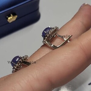 May include: A pair of silver earrings with a dark blue oval gemstone surrounded by small, clear stones. The earrings are held up by a person's fingers. The text "Vamika Jewels" is visible on the earrings.