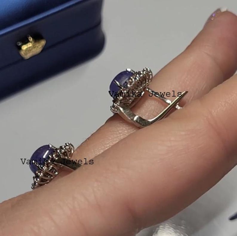 May include: A pair of silver earrings with a dark blue oval gemstone surrounded by small, clear stones. The earrings are held up by a person's fingers. The text "Vamika Jewels" is visible on the earrings.