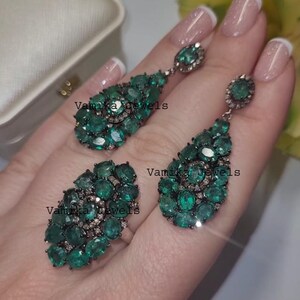 May include: A jewellery set comprising a ring and earrings. The ring and earrings are embellished with multiple oval-cut emerald green gemstones. The earrings are teardrop-shaped. The text "Vamika Jewels" is visible on the jewellery.