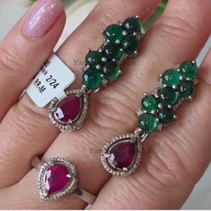 May include: A silver ring and earrings set. The ring features a teardrop-shaped ruby surrounded by small diamonds. The earrings have a ruby teardrop and a cluster of green stones. The jewelry is set in silver.