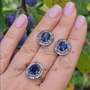 May include: A set of three silver rings featuring a large oval blue gemstone encircled by a halo of smaller multi-coloured gemstones. The rings are displayed on a hand with pink nail polish.