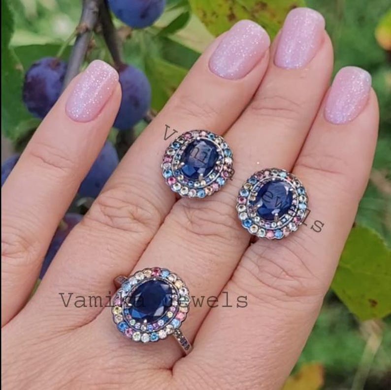 May include: A set of three silver rings featuring a large oval blue gemstone encircled by a halo of smaller multi-coloured gemstones. The rings are displayed on a hand with pink nail polish.