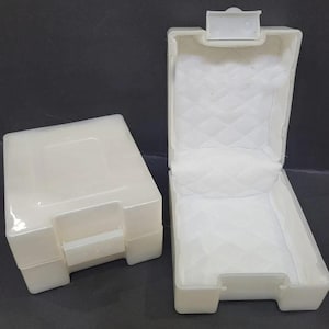 Pu&ograve; includere: Due scatole di plastica bianco sporco, una chiusa e una aperta. La scatola aperta ha un interno bianco trapuntato. Entrambe le scatole hanno una forma rettangolare con chiusura a scatto. Le scatole sono probabilmente per la conservazione o l'esposizione di piccoli oggetti.