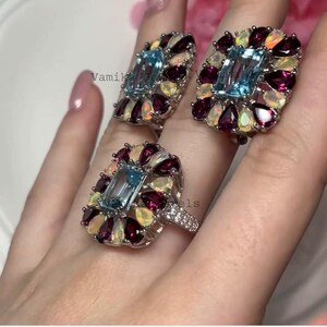 May include: A set of three gemstone rings and earrings. Each piece features a rectangular, light blue gemstone surrounded by smaller, colorful gems in a floral arrangement. The rings and earrings are set in silver.